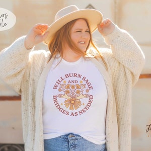 May include: White t-shirt featuring a graphic design of a hand holding a bouquet of flowers with the text "Will Burn Sage and Bridges as Needed".