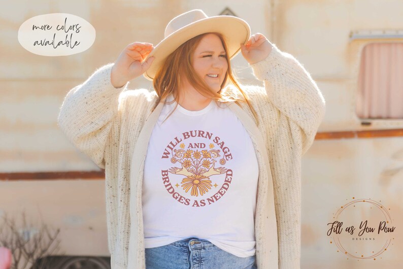 May include: White t-shirt featuring a graphic design of a hand holding a bouquet of flowers with the text "Will Burn Sage and Bridges as Needed".