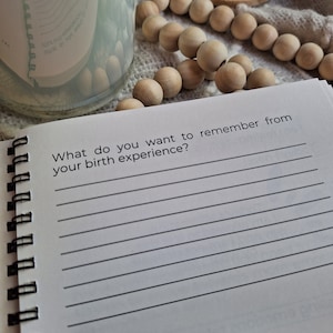 May include: A white journal with a black spiral binding. The journal has a lined page with the text "What do you want to remember from your birth experience?" and a blank page with lines for writing. The journal is open to a page with the text "See 3 what's around us, with that all life is".