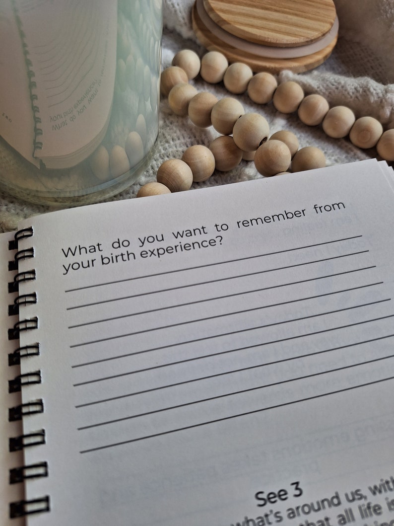 May include: A white journal with a black spiral binding. The journal has a lined page with the text "What do you want to remember from your birth experience?" and a blank page with lines for writing. The journal is open to a page with the text "See 3 what's around us, with that all life is".