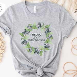 Op de afbeelding: Een grijs T-shirt met een krans van vlierbessen en de tekst "respecteer je vlierbessen".