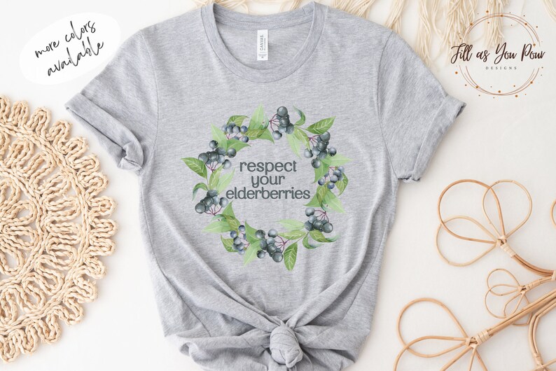 Op de afbeelding: Een grijs T-shirt met een krans van vlierbessen en de tekst "respecteer je vlierbessen".