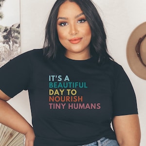 Puede incluir: Camiseta negra con el texto "It's a beautiful day to nourish tiny humans" en turquesa, azul, naranja y rosa. La camiseta tiene cuello redondo y mangas cortas. Un sombrero beige cuelga en la pared en el fondo.