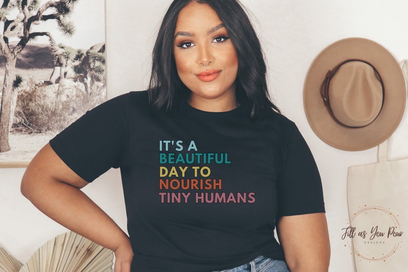 Puede incluir: Camiseta negra con el texto "It's a beautiful day to nourish tiny humans" en turquesa, azul, naranja y rosa. La camiseta tiene cuello redondo y mangas cortas. Un sombrero beige cuelga en la pared en el fondo.