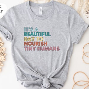 Puede incluir: Camiseta gris claro con el texto "IT'S A BEAUTIFUL DAY TO NOURISH TINY HUMANS" en letras de colores. La camiseta tiene mangas enrolladas y cuello redondo. Las palabras "more colors available" est&aacute;n en la esquina superior izquierda.