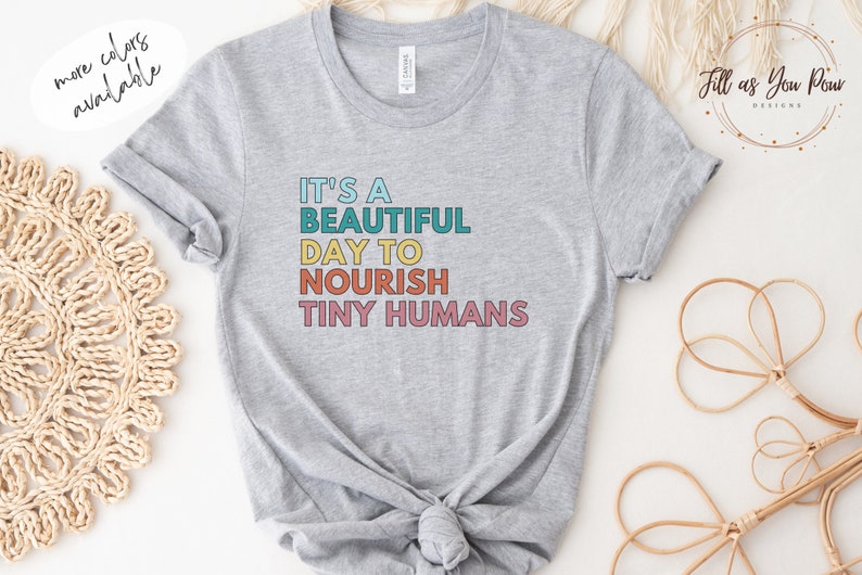Puede incluir: Camiseta gris claro con el texto "IT'S A BEAUTIFUL DAY TO NOURISH TINY HUMANS" en letras de colores. La camiseta tiene mangas enrolladas y cuello redondo. Las palabras "more colors available" est&aacute;n en la esquina superior izquierda.