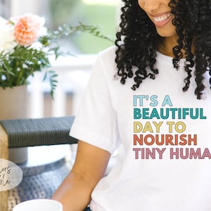 Puede incluir: Camiseta blanca con el texto "IT'S A BEAUTIFUL DAY TO NOURISH TINY HUMANS" en letras coloridas. La camiseta la lleva una persona con el pelo rizado. Un jarr&oacute;n con flores est&aacute; en el fondo.