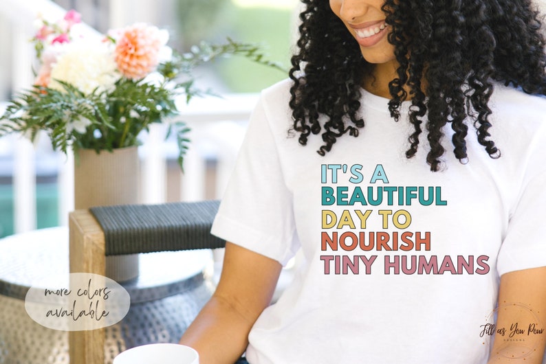 Puede incluir: Camiseta blanca con el texto "IT'S A BEAUTIFUL DAY TO NOURISH TINY HUMANS" en letras coloridas. La camiseta la lleva una persona con el pelo rizado. Un jarr&oacute;n con flores est&aacute; en el fondo.