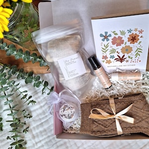 Puede incluir: Una caja de regalo llena de art&iacute;culos de cuidado personal, que incluyen sales de ba&ntilde;o, aceite esencial, b&aacute;lsamo labial, calcetines y una bomba de ba&ntilde;o. La caja est&aacute; decorada con papel marr&oacute;n y papel de seda blanco. La tarjeta dice "Madre como ninguna otra".