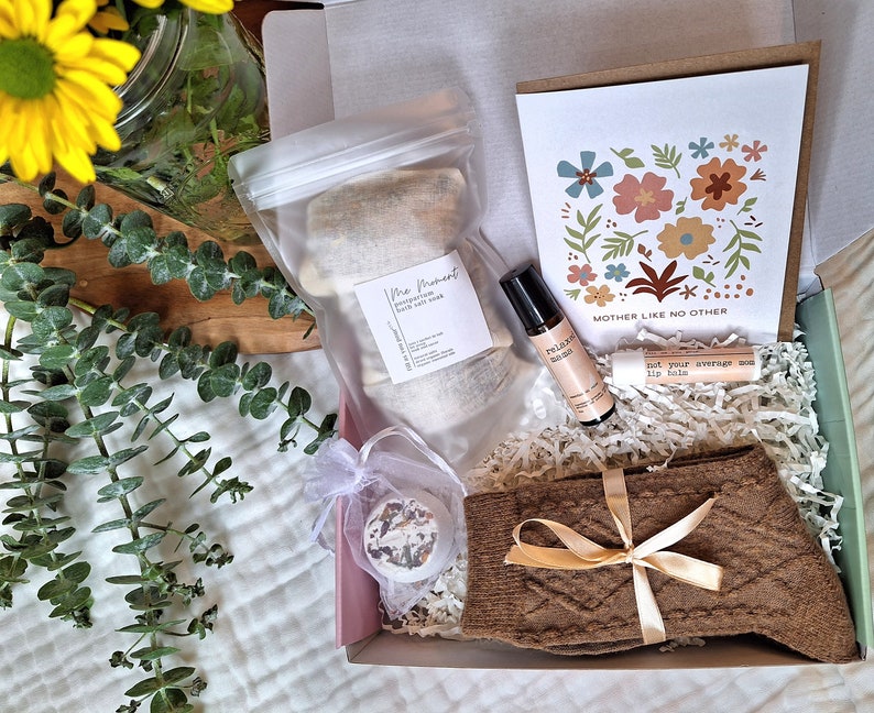 Puede incluir: Una caja de regalo llena de art&iacute;culos de cuidado personal, que incluyen sales de ba&ntilde;o, aceite esencial, b&aacute;lsamo labial, calcetines y una bomba de ba&ntilde;o. La caja est&aacute; decorada con papel marr&oacute;n y papel de seda blanco. La tarjeta dice "Madre como ninguna otra".