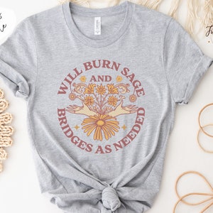 May include: Grey t-shirt featuring a graphic design that reads "Will Burn Sage and Bridges as Needed". The graphic depicts a bouquet of flowers with two hands reaching out from the sides.