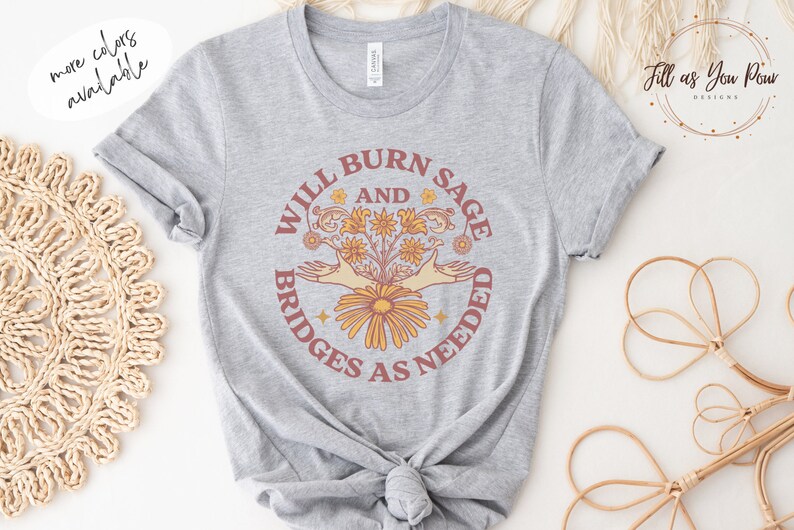 May include: Grey t-shirt featuring a graphic design that reads "Will Burn Sage and Bridges as Needed". The graphic depicts a bouquet of flowers with two hands reaching out from the sides.
