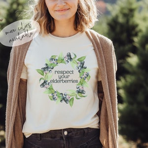 Op de afbeelding: Een wit T-shirt met een aquarelontwerp van vlierbessen en de tekst "respecteer je vlierbessen".