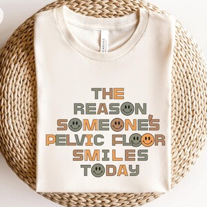 Puede incluir: Una camiseta beige con el texto "THE REASON SOMEONE'S PELVIC FLOOR SMILES TODAY" en letras marrones, naranjas y verdes. El texto está rodeado de caritas sonrientes de varios colores.