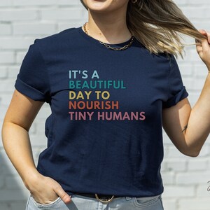 Puede incluir: Camiseta azul marino con el texto "IT'S A BEAUTIFUL DAY TO NOURISH TINY HUMANS" en verde azulado, amarillo, naranja y rosa. La camiseta es de cuello redondo y tiene las mangas enrolladas. La camiseta la lleva una persona.