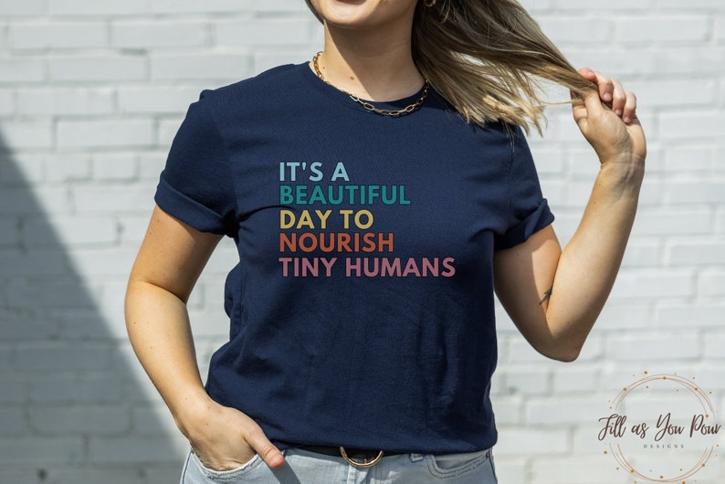 Puede incluir: Camiseta azul marino con el texto "IT'S A BEAUTIFUL DAY TO NOURISH TINY HUMANS" en verde azulado, amarillo, naranja y rosa. La camiseta es de cuello redondo y tiene las mangas enrolladas. La camiseta la lleva una persona.