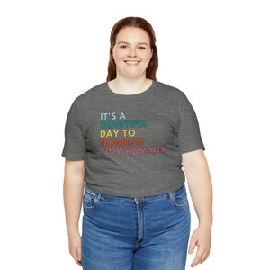 Puede incluir: Una camiseta gris con el texto "IT'S A BEAUTIFUL DAY TO NOURISH TINY HUMANS" en turquesa, azul, naranja y rosa. La camiseta se combina con unos vaqueros azules.