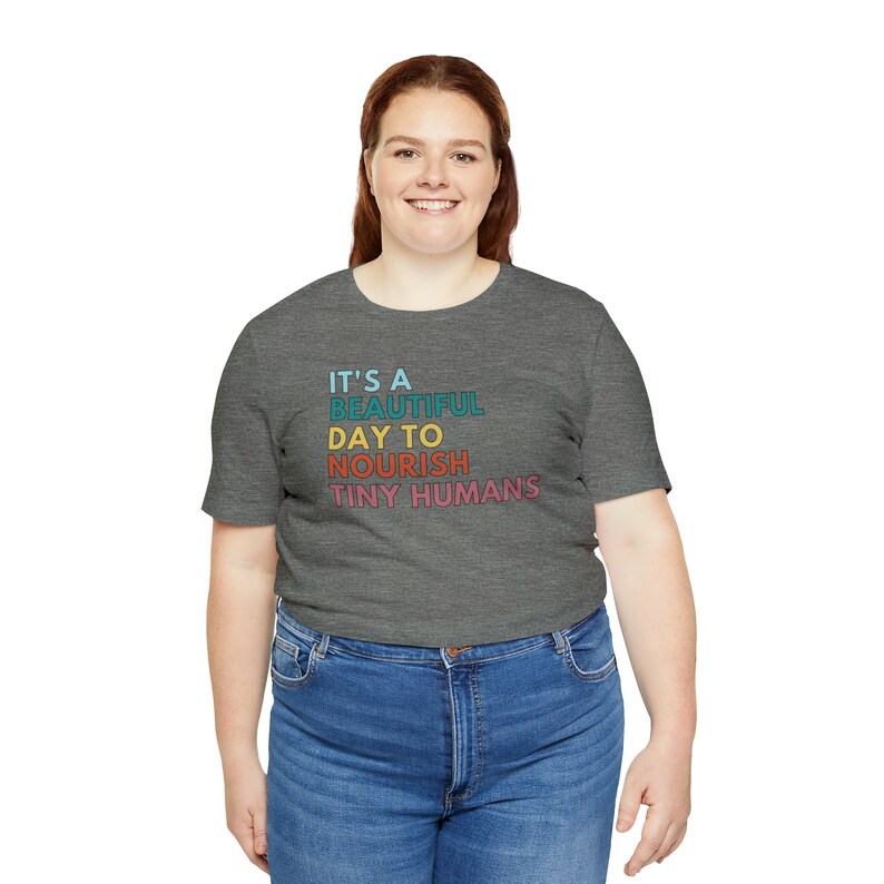 Puede incluir: Una camiseta gris con el texto "IT'S A BEAUTIFUL DAY TO NOURISH TINY HUMANS" en turquesa, azul, naranja y rosa. La camiseta se combina con unos vaqueros azules.