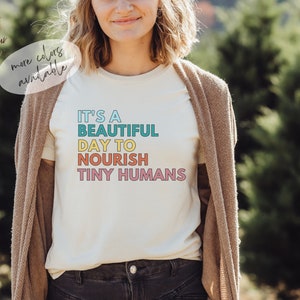 Puede incluir: Camiseta color crema con el texto "IT'S A BEAUTIFUL DAY TO NOURISH TINY HUMANS" en turquesa, amarillo, naranja y rosa. La persona lleva una rebeca marr&oacute;n. M&aacute;s colores disponibles.