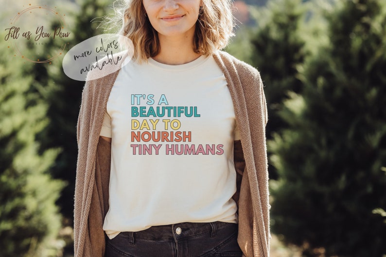 Puede incluir: Camiseta color crema con el texto "IT'S A BEAUTIFUL DAY TO NOURISH TINY HUMANS" en turquesa, amarillo, naranja y rosa. La persona lleva una rebeca marr&oacute;n. M&aacute;s colores disponibles.