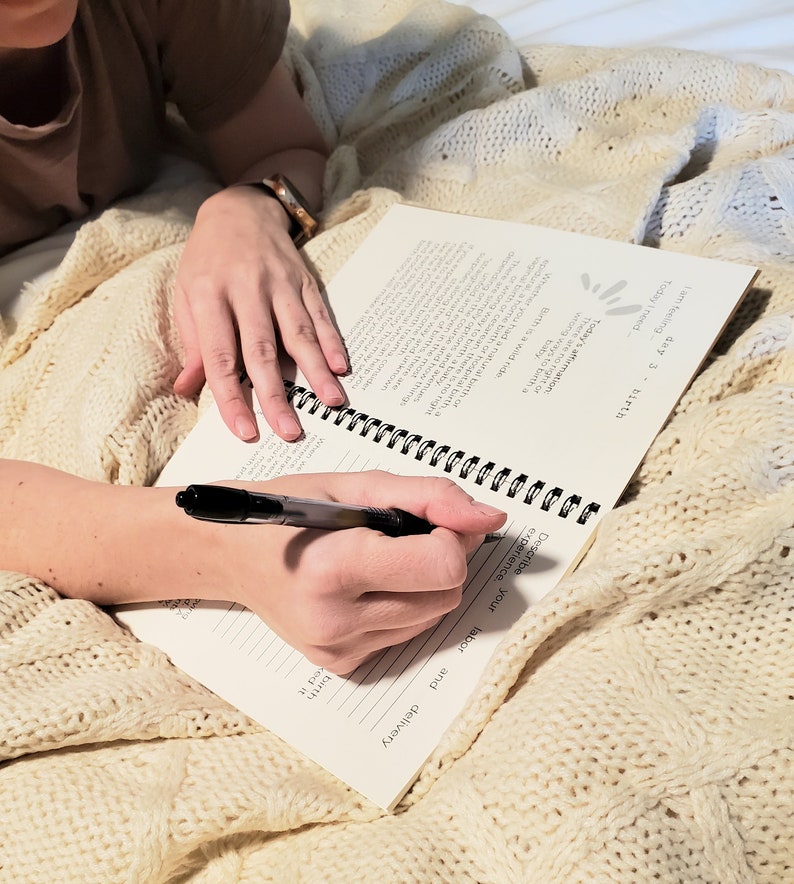 May include: A person writing in a spiral bound journal with a black pen. The journal has the text "day 3" and "birth" on the cover. The journal is open to a page with the text "Describe your labor and delivery".