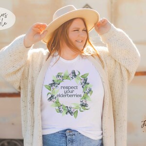 Op de afbeelding: Wit T-shirt met een aquarelkrans van vlierbessen en de tekst "respecteer je vlierbessen".