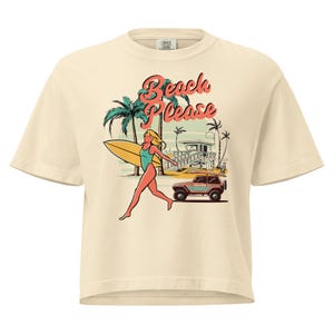 May include: Cream-colored t-shirt with a graphic featuring a surfer girl, palm trees, a beach shack, and a vintage SUV. The text "Beach Please" is displayed in a retro font. The design evokes a summery, beach-themed vibe.