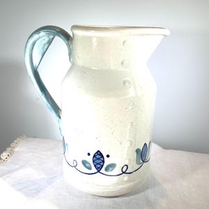 May include: A white ceramic pitcher with a blue handle and a blue floral design around the bottom. The pitcher has a speckled finish.