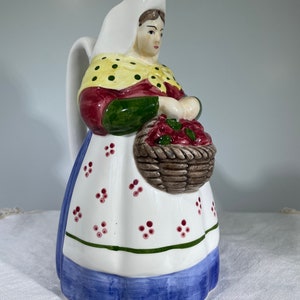 May include: A ceramic pitcher shaped like a woman in a white bonnet and blue and white dress holding a basket of red fruit. The pitcher has a yellow and green floral pattern.