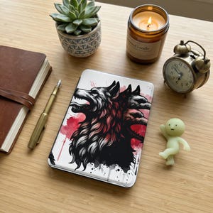 May include: A white phone case featuring a black and red illustration of a three-headed wolf. The case is on a wooden surface, next to a candle, a succulent, a small clock, a pen, a notebook, and a small figurine.