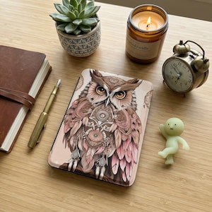 Puede incluir: Una funda de teléfono decorativa con una ilustración detallada de un búho en tonos marrón, rosa y beige. La funda está rodeada de una planta suculenta, una vela encendida, un despertador vintage, una pequeña figura, un diario y un bolígrafo sobre una superficie de madera.