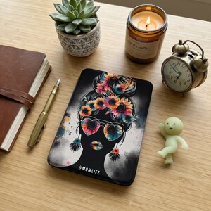 Può includere: Una custodia per telefono nera con un design floreale raffigurante la silhouette di una donna con occhiali da sole a forma di cuore e una corona di fiori. La custodia ha il testo "#MOMLIFE" in basso. Altri oggetti includono una candela, un orologio, una succulenta e una piccola statuina.
