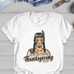 Puede incluir: Camiseta blanca con una ilustración de dibujos animados de una mujer nativa americana con un tocado y un atuendo verde y marrón. El texto "You're welcome... Thanksgiving 1621" está impreso debajo de la ilustración.
