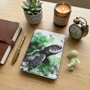 May include: A white and green phone case featuring a detailed illustration of a sea turtle. The design incorporates watercolor-style green splashes. Other items include a succulent, a candle, a vintage alarm clock, a pen, a notebook, and a small figurine.