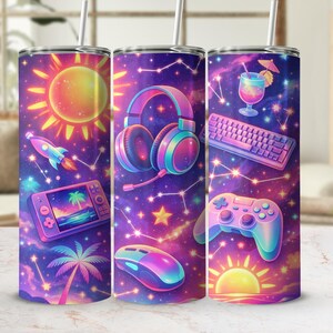 Puede incluir: Tres vasos de acero inoxidable con un diseño vibrante y colorido. Cada vaso presenta una ilustración diferente con temática de juegos sobre un fondo estrellado, similar a una galaxia. Los diseños incluyen un sol, un cohete, auriculares, un teclado y un mando.
