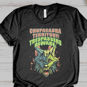 Pode incluir: Camiseta preta com um gráfico de uma criatura semelhante a um lobo rosnando. O texto "CHUPACABRA TERRITORY! TRESPASSERS BEWARE" está impresso acima da criatura em laranja e verde. O design tem um visual vintage.