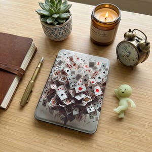 May include: A rectangular playing card design on a white surface. The design features playing cards with red, black, and white suits. A small succulent plant, a candle, a vintage clock, a notebook, a pen, and a small figurine are also present.