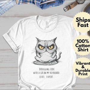 Pode incluir: Camiseta branca com uma ilustração em preto e branco de um gato sentado em um teclado. O texto na camiseta diz "Debugging code with a cat on my keyboard. Level Expert."