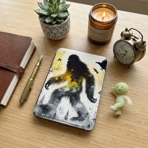 Può includere: Custodia per telefono bianca con un design Bigfoot nero e giallo. Su un tavolo di legno, accanto a una succulenta, una candela accesa, una piccola sveglia, una statuina, un diario e una penna.
