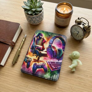 May include: A gaming-themed mousepad with a colorful design featuring a gaming chair, headphones, keyboard, and controllers. The mousepad is surrounded by a succulent plant, a lit candle, a vintage alarm clock, a pen, a notebook, and a small figurine.