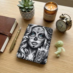 May include: A black and white decorative phone case featuring a woman with sugar skull makeup and blue eyes. The case has intricate floral and spiderweb designs. A lit candle, succulent, and small clock are also on the wooden surface.