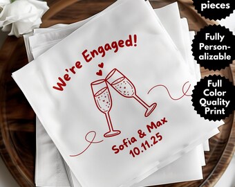 May include: White paper napkins with red text and a champagne glass design. The napkins read "We're Engaged!" with the names "Sofia & Max" and the date "10.11.25". The napkins are stacked on a wooden tray.