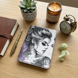 以下が含まれることがあります： タトゥーと蝶の女性の白黒イラストが描かれた装飾的なジャーナル。紫色の水彩効果があります。ジャーナルは木製の表面にあり、多肉植物、キャンドル、ヴィンテージ時計、小さなフィギュアが並んでいます。
