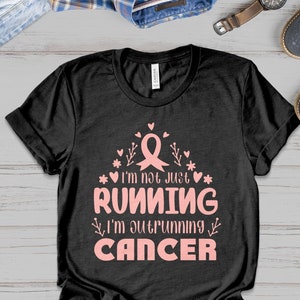 Pode incluir: Camiseta preta com uma fita rosa, corações e o texto "Não estou apenas correndo, estou superando o câncer".