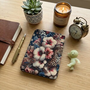 Può includere: Una custodia per tablet con un motivo floreale di fiori di ibisco e un motivo leopardato. La custodia è circondata da una pianta succulenta, una candela accesa, una sveglia vintage, una piccola statuina, una penna e un diario rilegato in pelle.