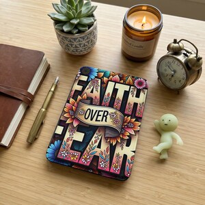 Può includere: Un taccuino decorativo con le parole "FAITH OVER FEAR" in un design floreale, un diario in pelle marrone, una penna d'oro, una pianta succulenta, una candela accesa, una sveglia vintage e una piccola statuina sono disposti su una superficie di legno.