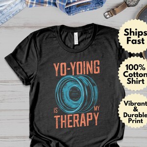 Może przedstawiać: Czarna koszulka z napisem "YO-YOING IS MY THERAPY" w kolorze pomarańczowym, z niebieską grafiką jo-jo. Koszulka jest wykonana w 100% z bawełny i ma żywy i trwały nadruk.