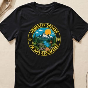 Może przedstawiać: Czarny t-shirt z krótkim rękawem z okrągłą grafiką przedstawiającą górski krajobraz, rzekę, pojemnik do geocachingu i słońce. Napis HONESTLY OFFICER I'M JUST GEOCACHING widnieje żółtymi literami wokół krawędzi okrągłego wzoru.