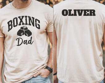 May include: Beige t-shirt with the text "BOXING Dad" above a pair of boxing gloves in black. The back of the shirt displays the name "OLIVER" in bold, black letters. The shirt is short-sleeved.