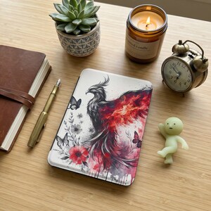 May include: A white tablet case featuring a phoenix design in shades of red, black, and white, with butterflies and flowers. A small, vintage-style alarm clock, a lit candle, and a succulent plant are also visible.